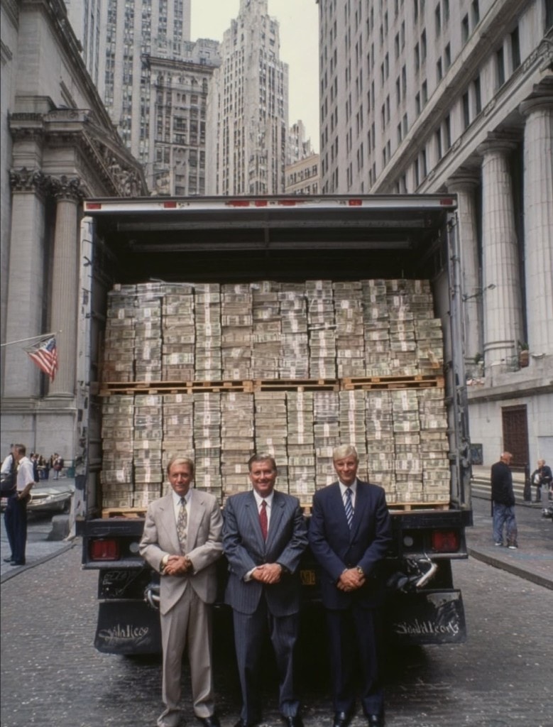Three men in suits standing in front of a truck packed with palletized cash.